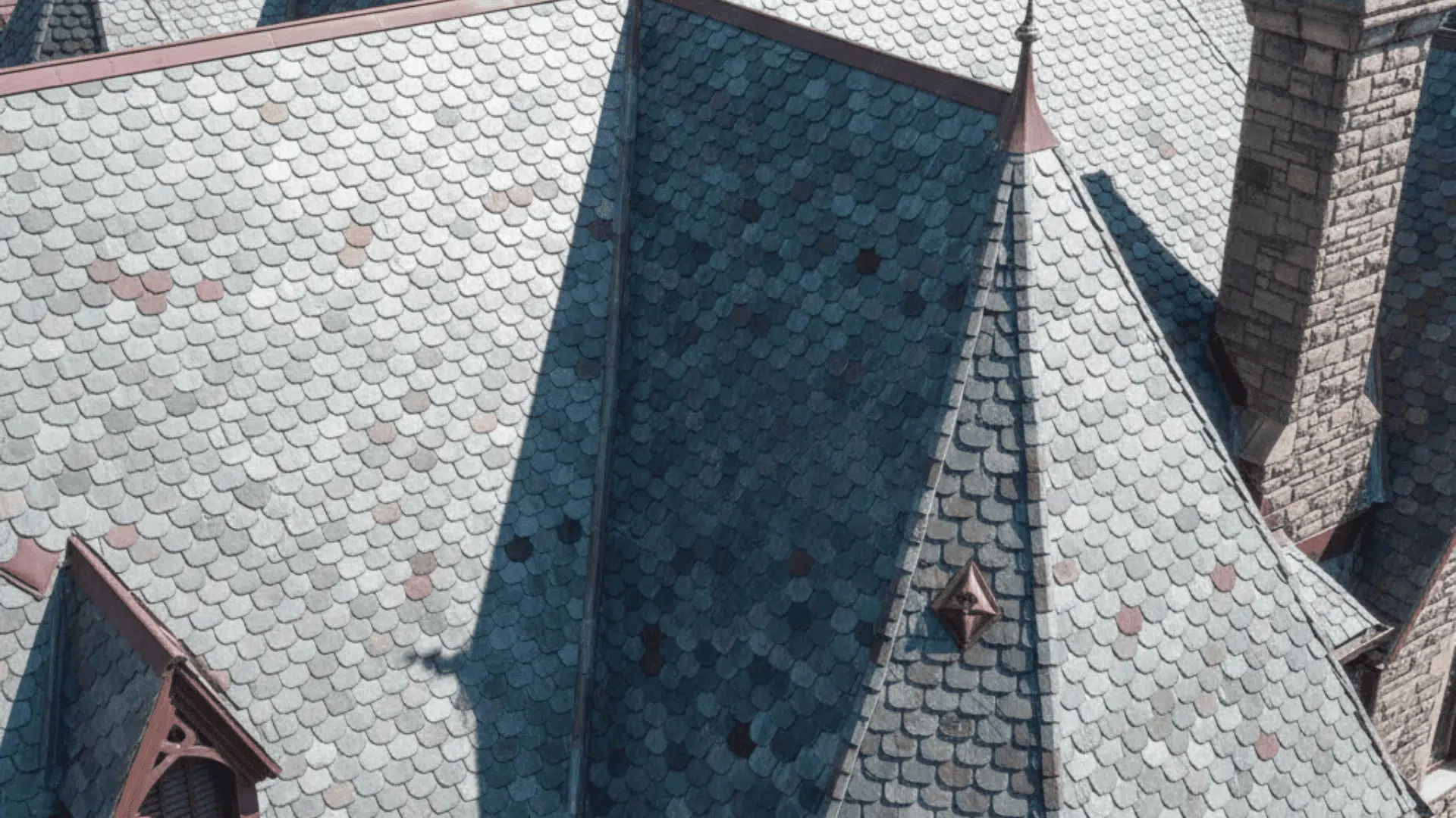 Top of a gothic building with a cross, featuring a decorative slate roof in various colors arranged in diamond patterns.