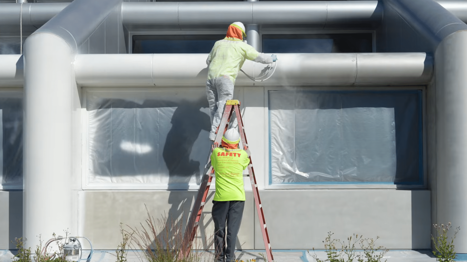Worker on a ladder wearing protective gear while spraying an exterior metal surface, demonstrating a safe setup before painting.