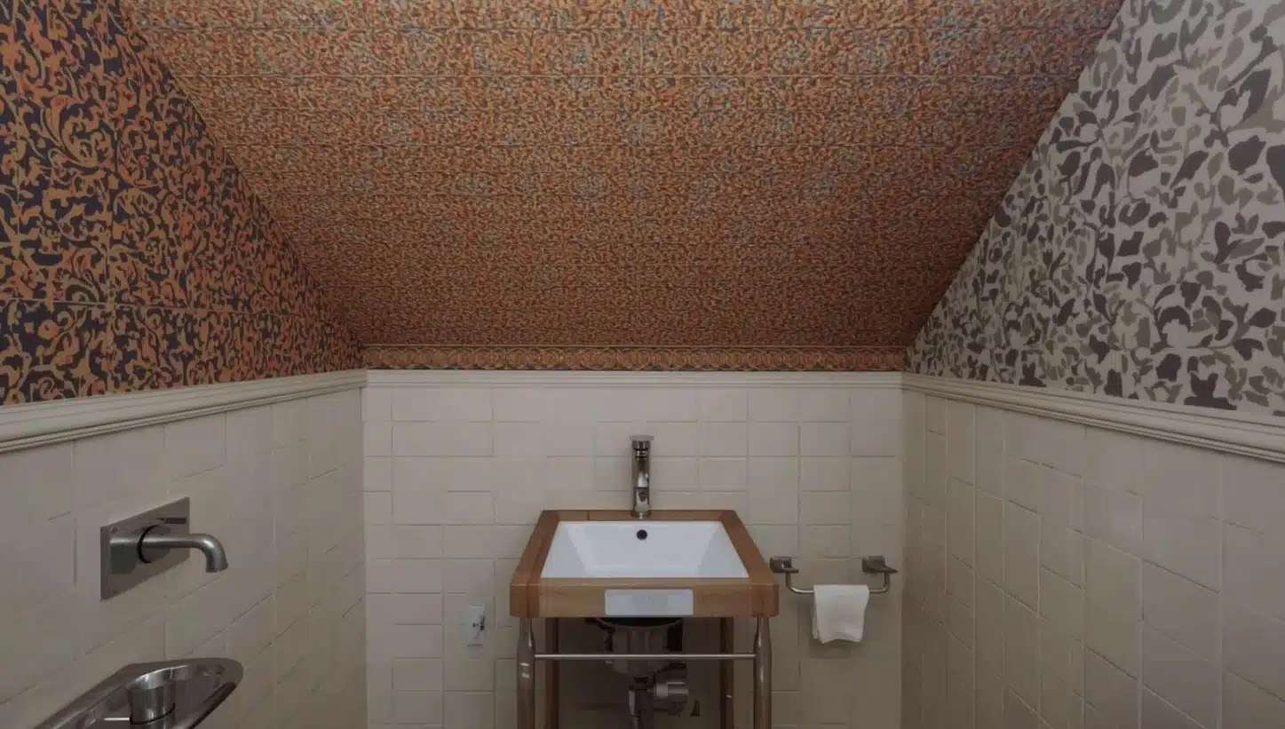 Cozy powder bath with white tile wainscoting and bold patterned wallpaper covering sloped ceiling and upper walls.